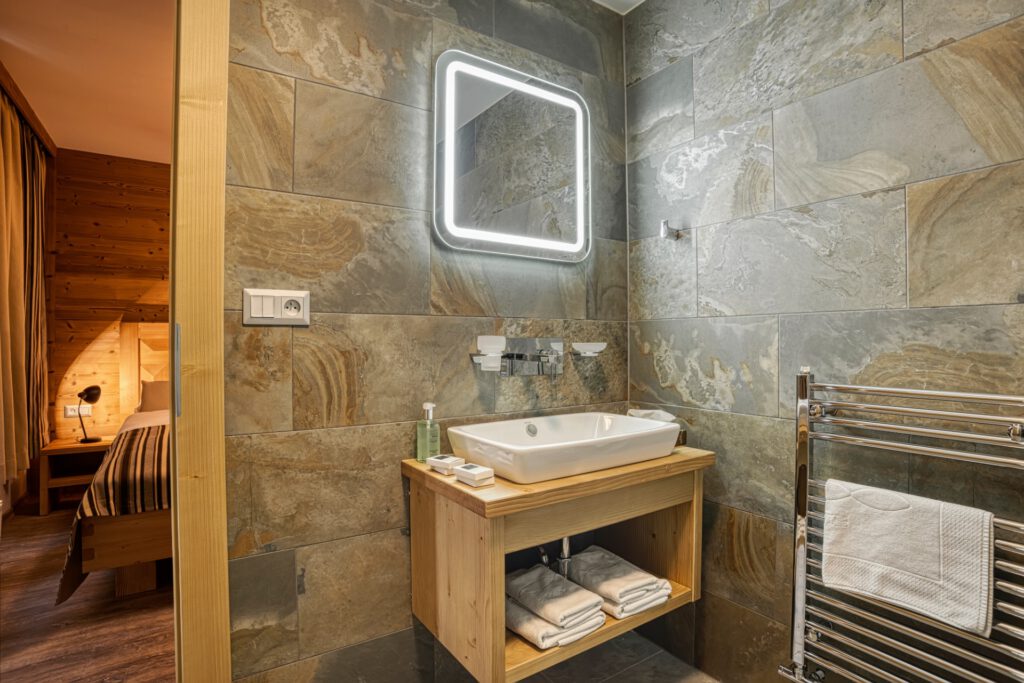 Moderná kúpeľňa v apartmáne Gabor Apartments s kamenným obkladom, dizajnovým umývadlom, osvetleným zrkadlom a drevenými prvkami.
